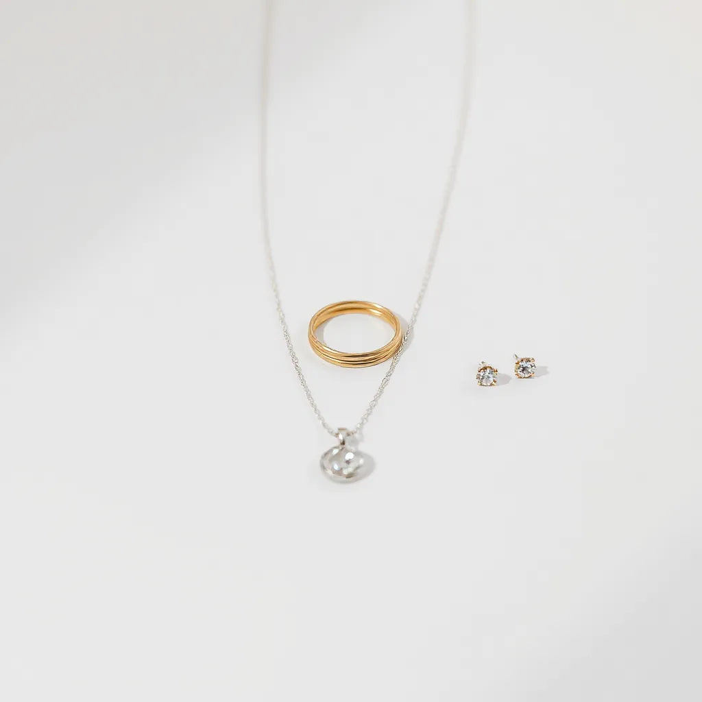 Delicate minimalist jewelry set with rings, pendant necklace, and stud earrings on a light neutral surface.